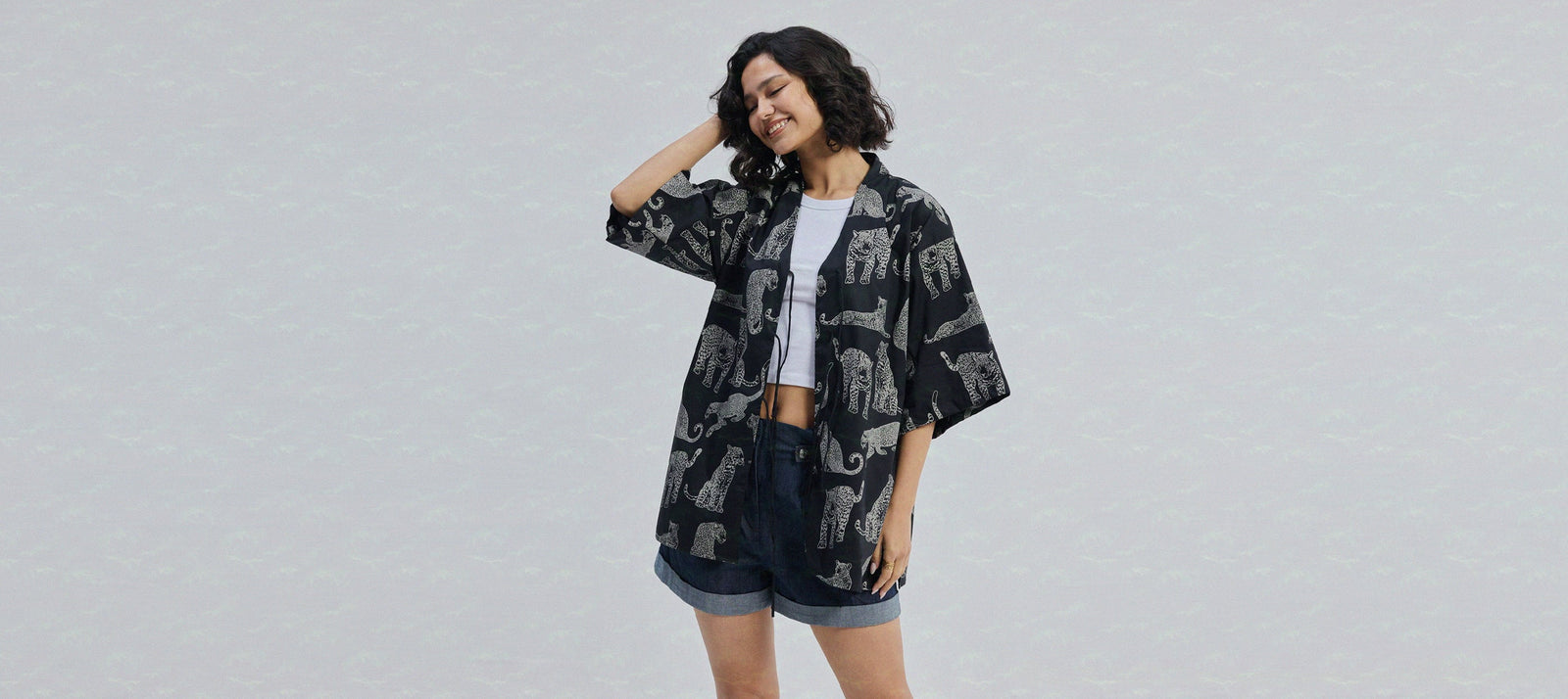 Woman wearing a black floral kimono jacket over a white crop top and shorts, posing against a grey studio background.