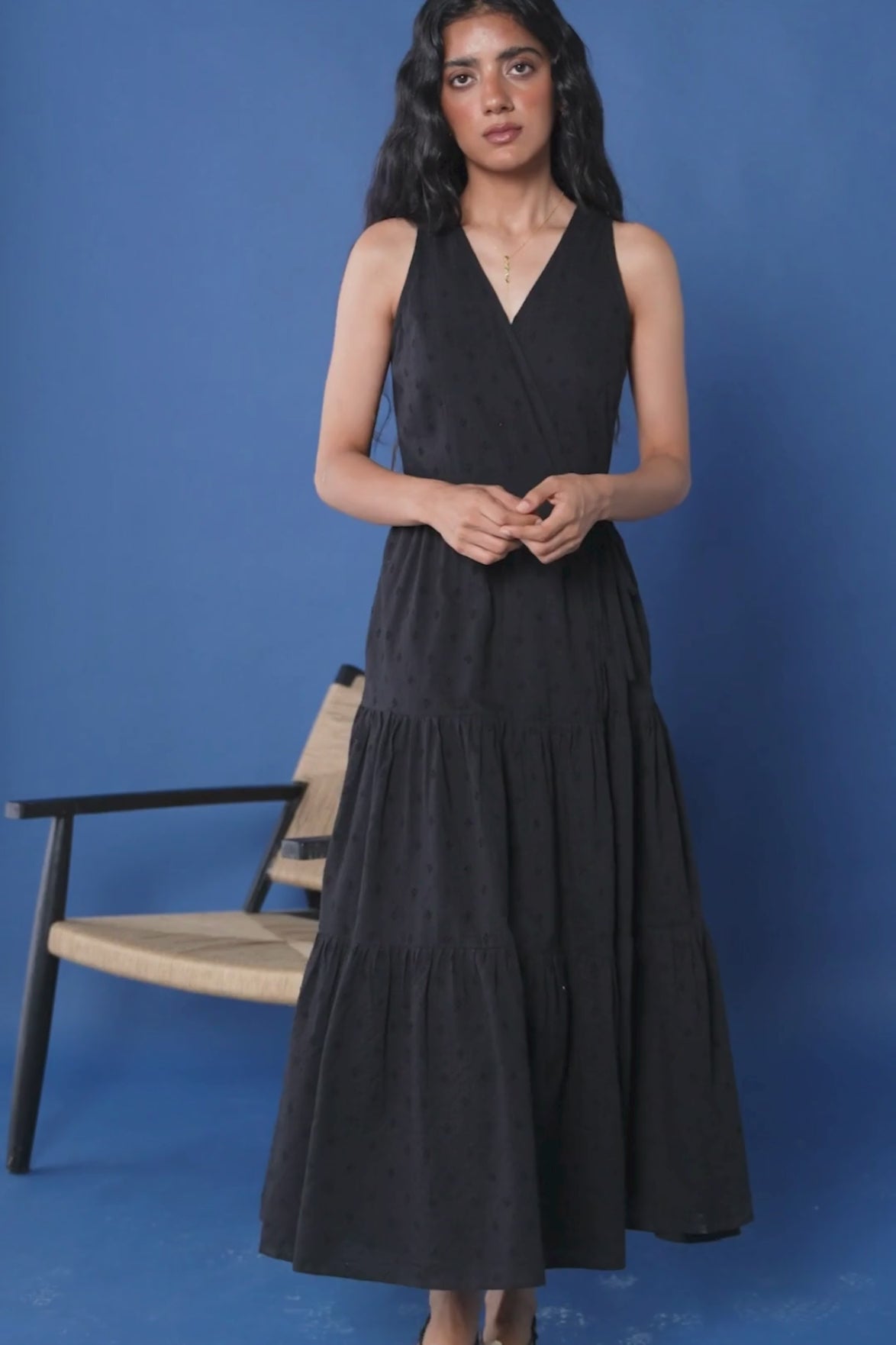 Model presents the Sleeveless Embroidered Wrap Dress with Tiers in Black, walking gracefully, completing a soft twirl to display the tiered design, and taking a seat on a chair to highlight the fabric movement and elegant drape.